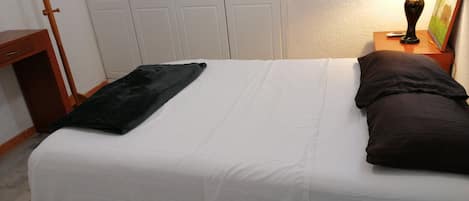Un lit bien fait, avec du linge de lit blanc et un plaid noir, un porte-manteau en bois et une lampe sur une table de chevet.