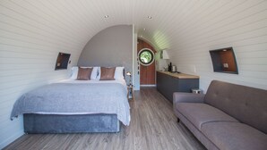 A modern bedroom with a large bed, a sofa, and a kitchen area with a round window. - Primrose Family Glamping Pod | sleeps 4 - Gold Award winning (Pickering)