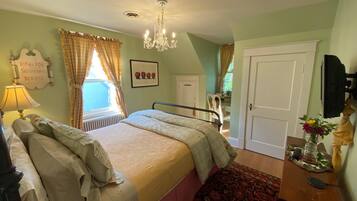 A bedroom with a bed, a bedside table, a lamp, a radiator, a window with curtains, a door, and a television.