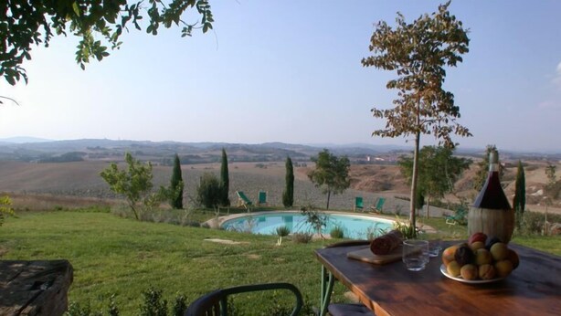 Une table placée au bord de la piscine, avec une bouteille, des verres et des fruits, d’où l’on a vue sur la piscine et la campagne environnante.