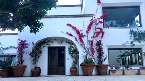 A white building with a large arched doorway, multiple potted plants with flowers, and a clear view of the sky. - Tenuta DonnAnna (Galatone)