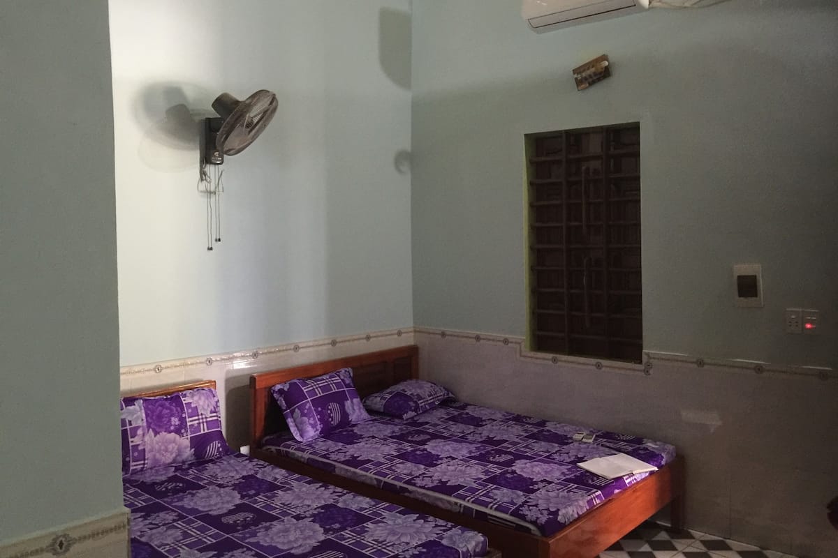Una habitación con dos camas, un ventilador de pared, un equipo de aire acondicionado y una ventana con diseño de celosía.