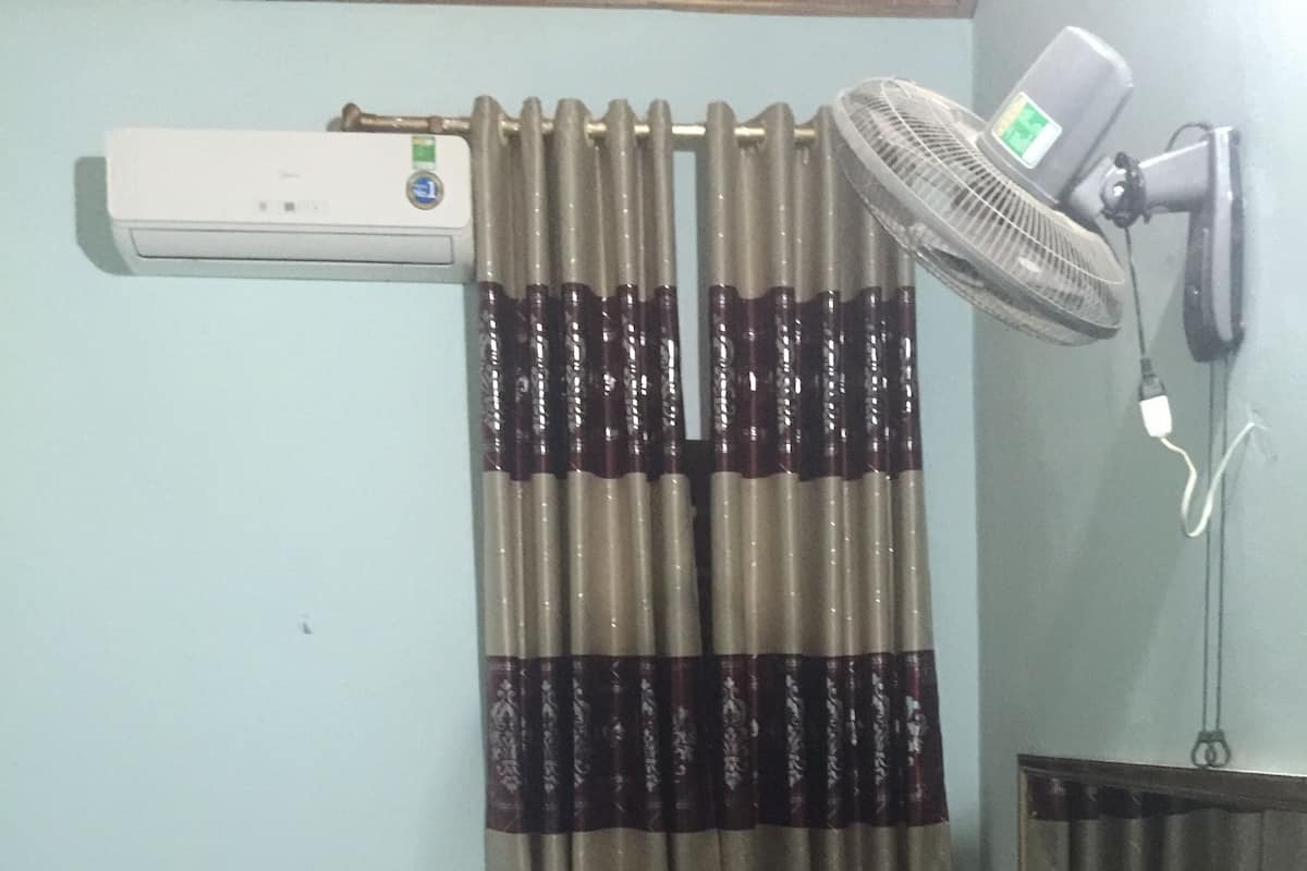 Una habitación con cama, escritorio, dos sillas, ventilador de pie, aire acondicionado y espejo.