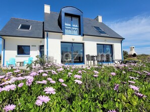 A modern house with a large glass door, a blue roof, and a garden with purple flowers. - Tipoz'Immo-Les Galets- sea view house - fully equipped - pets welcome (Plozévet)