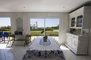 A dining area with a table set for two, a white cabinet with glass doors, and a view of a house and garden through a sliding door. - Tipoz'Immo-Les Galets- sea view house - fully equipped - pets welcome (Plozévet)