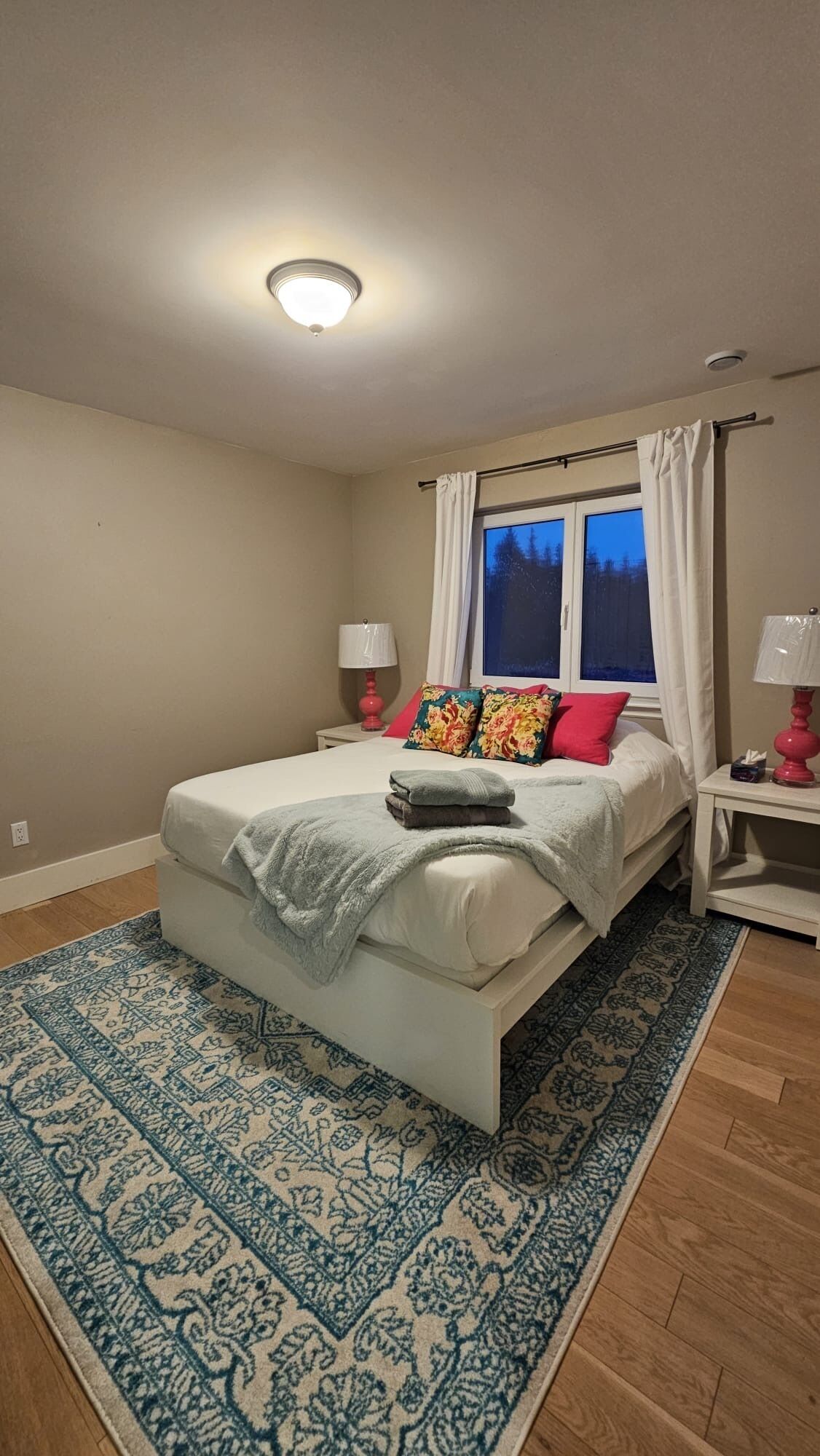 Une chambre à coucher avec un lit, une fenêtre avec des rideaux, une table de chevet avec une lampe et un tapis à motifs.