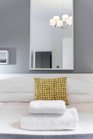 A modern hotel room with a large mirror, a chandelier, a white bed with a patterned pillow, and neatly folded towels.