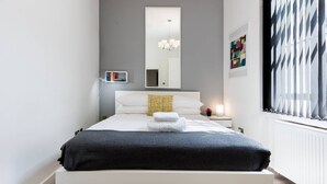 A modern bedroom with a bed, bedside tables, a nightstand, a mirror, and a window with blinds.