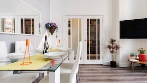 A modern dining area with a glass table, white chairs, and a bookshelf in the background.