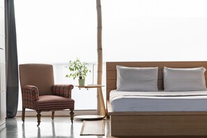 A bedroom with a bed, a chair, a small table with a plant, and a window with a view.