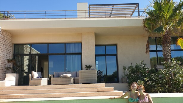 Un área moderna junto a la piscina con una piscina cristalina, mobiliario exterior y vistas a un edificio contemporáneo con amplias ventanas.