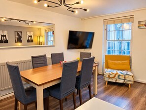 Un coin repas avec une table en bois et six chaises, un téléviseur à écran plat fixé au mur et une fenêtre avec des stores.