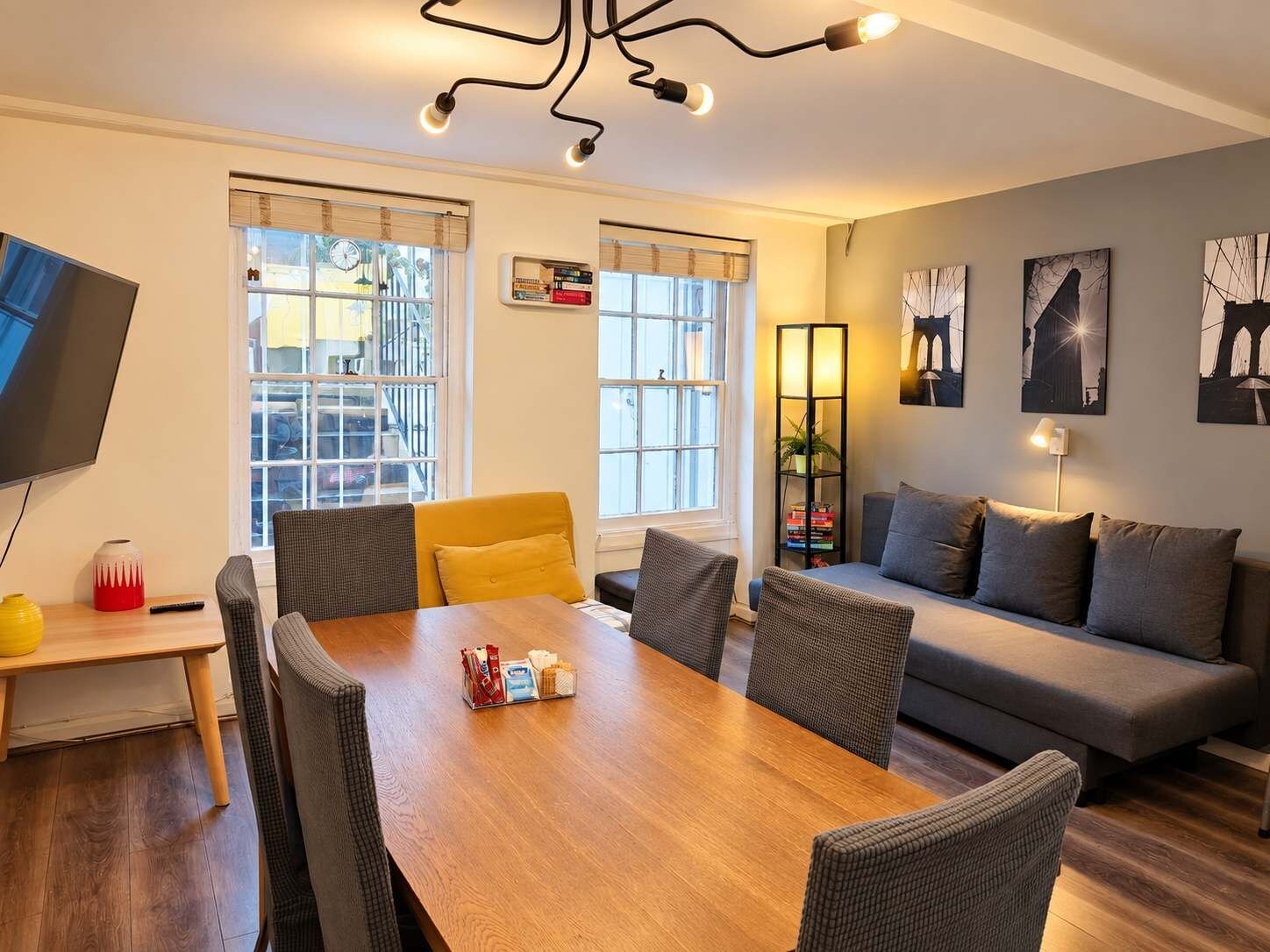 A modern living room with a wooden dining table, grey sofas, and a TV mounted on the wall.