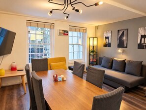 A modern living room with a wooden dining table, grey sofas, and a TV mounted on the wall.