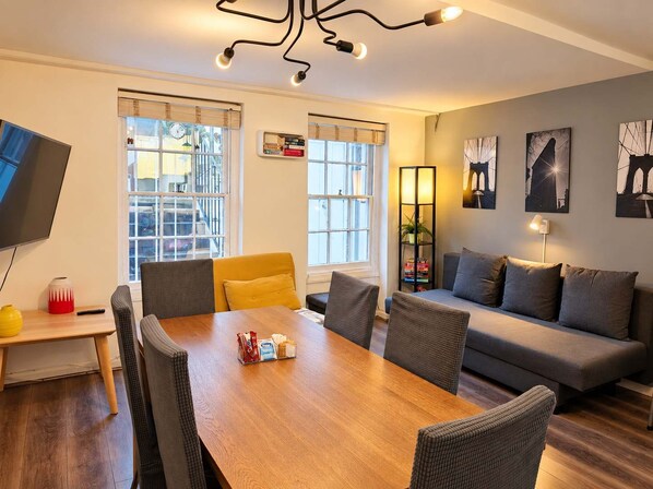 A modern living room with a wooden dining table, grey sofas, and a TV mounted on the wall.