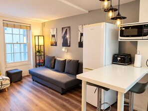 Un espace de vie compact comprenant un coin cuisine, un canapé, une table à manger et une fenêtre avec des stores.