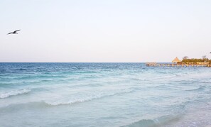 Ein Strand mit Wellen, einer Holzsteg und ein Gebäude mit einem Reetdach.