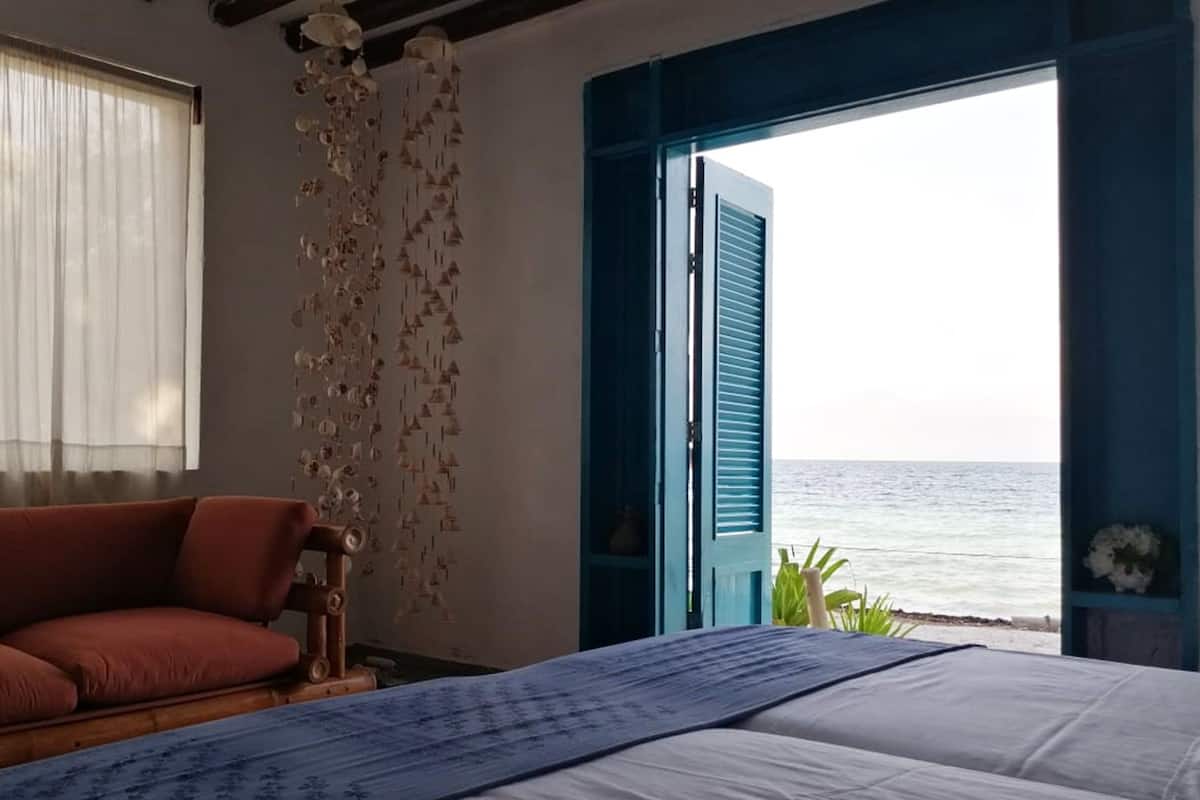 Una habitación con cama, un sofá rojo, una ventana con cortinas traslúcidas y vistas al mar mediante persianas azules.