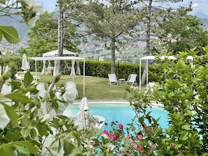 A poolside area with lounge chairs and a gazebo, surrounded by greenery and a view of distant hills.