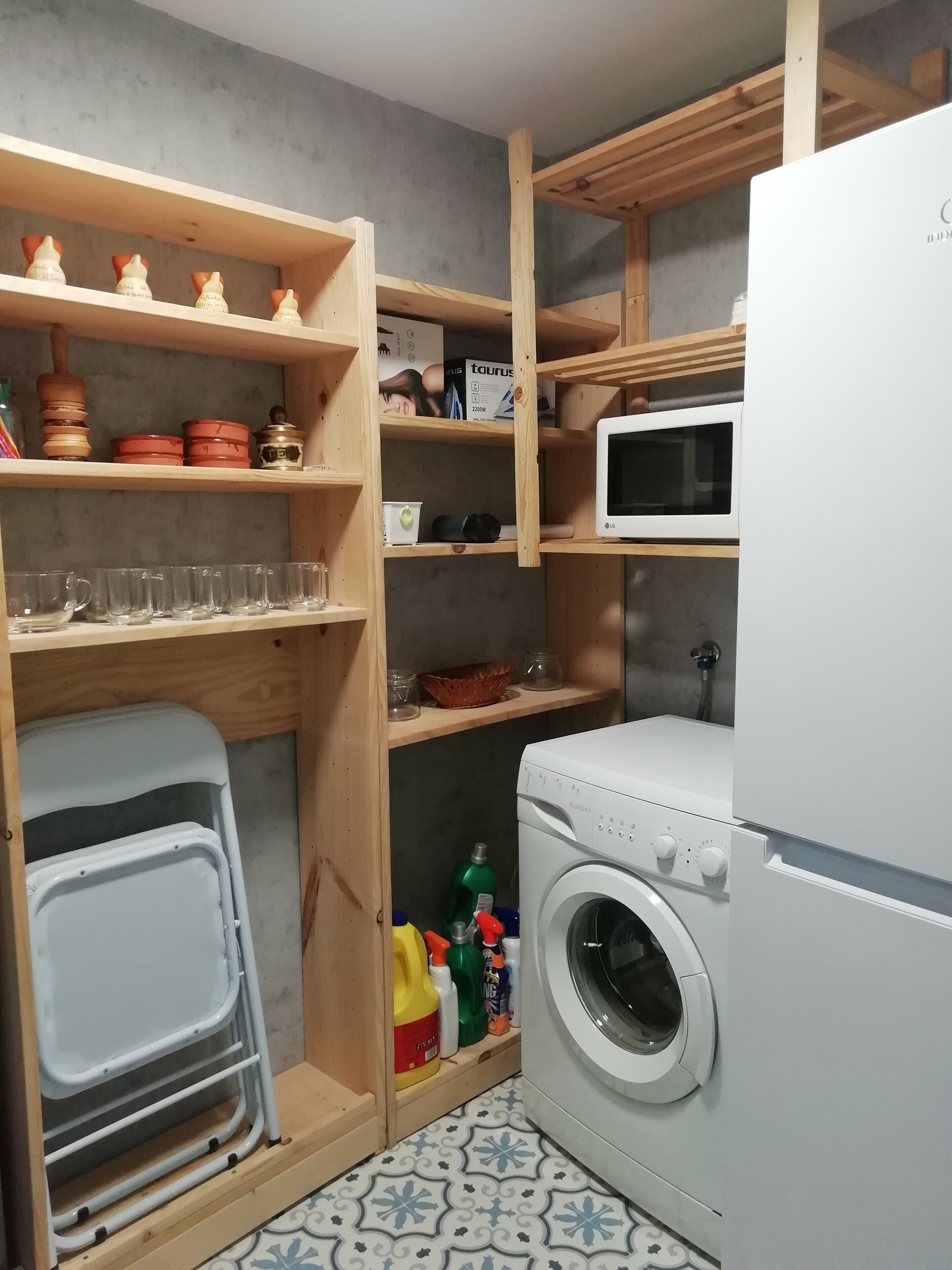 A laundry room with a washing machine, shelves with kitchenware, and a microwave.