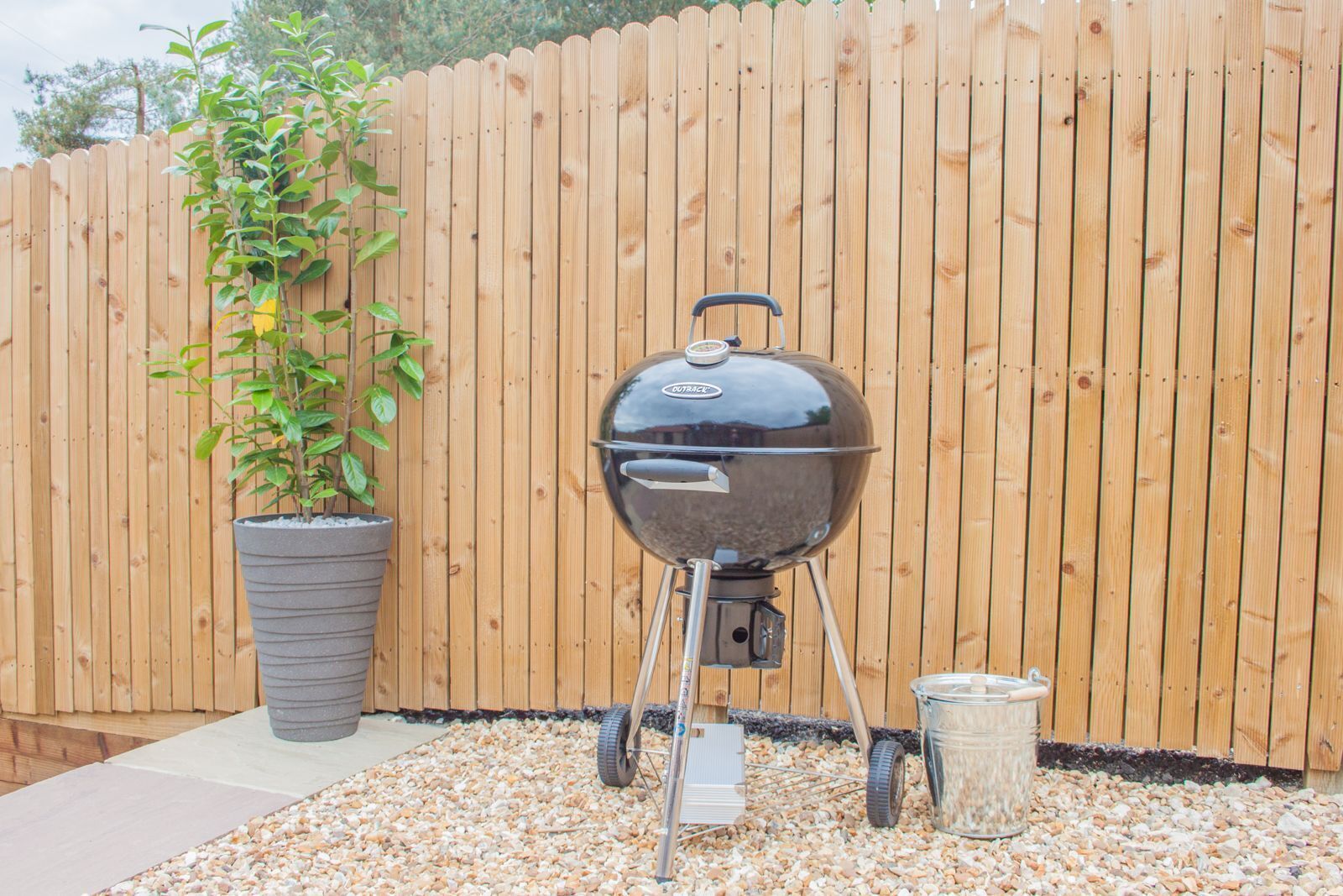 A barbecue grill with a pot of plants and a container of charcoal beside it.