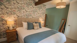 A bedroom with a large bed, two bedside tables with lamps, floral wallpaper, and a wooden ceiling beam.