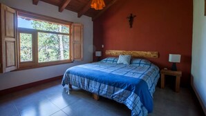 Une chambre avec un plafond en bois, un grand lit recouvert d’une couette à carreaux bleus et blancs, deux lampes de chevet et une fenêtre avec des volets en bois.
