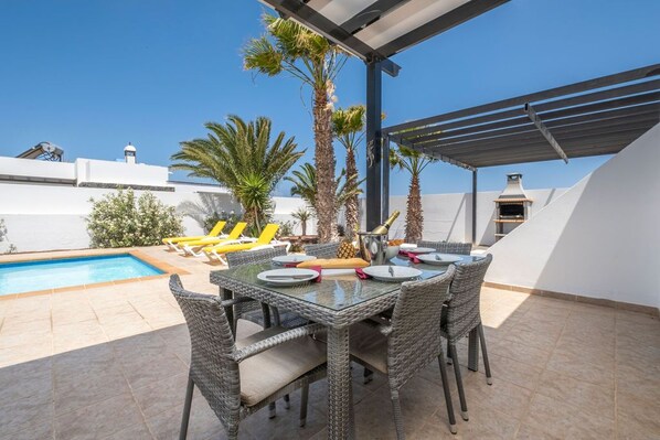 Un patio con tavolo in vetro, sedie in vimini e una piscina.