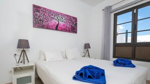 A hotel room with a bed, white pillows, a nightstand with a lamp, a painting of a tree, and a window with curtains. - Villa Dulce Vista - 3 bedrooms, Heated Private Pool, with Mountain Views (Playa Blanca)