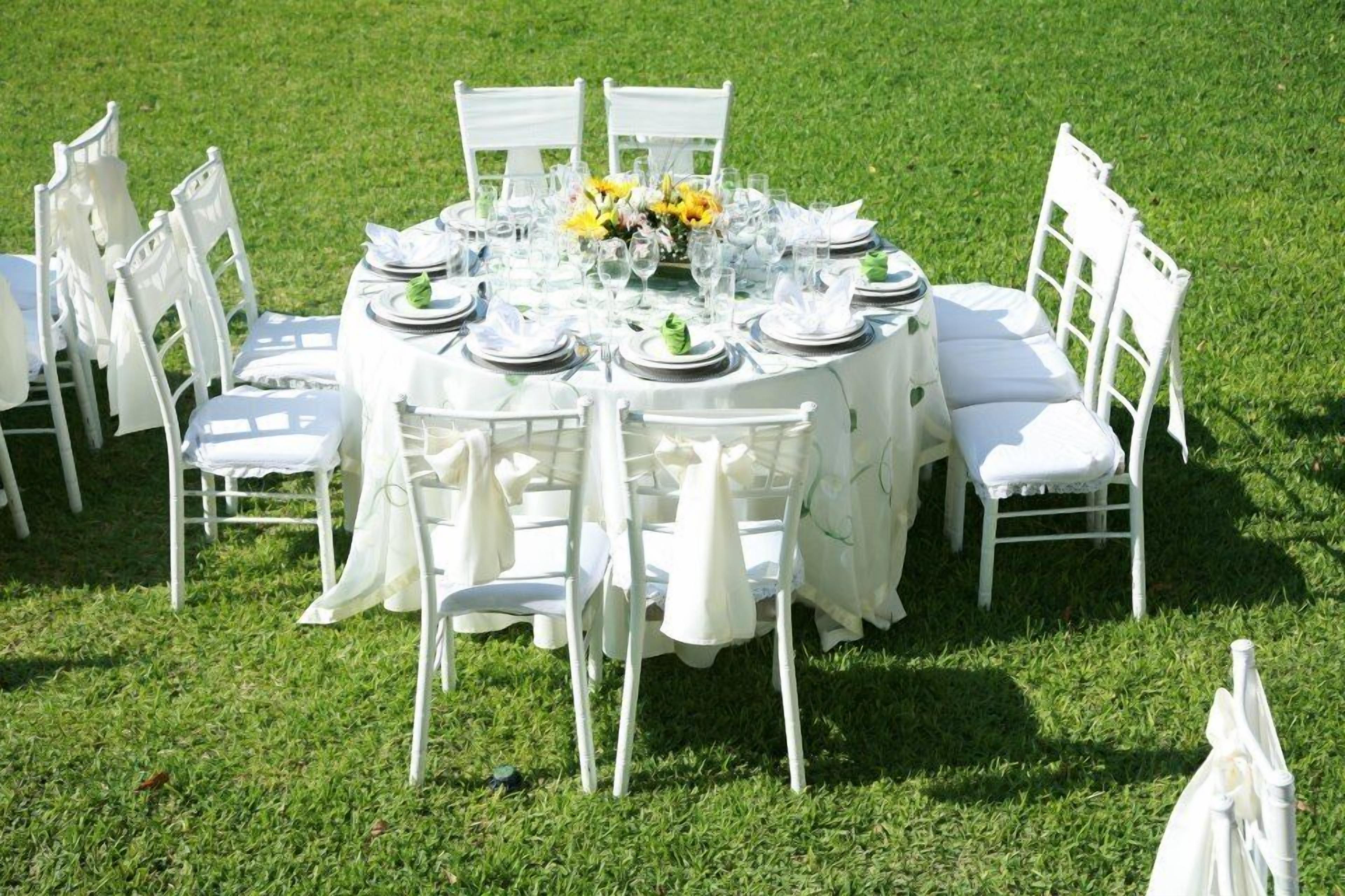 A round table set outdoors with white chairs, a floral centerpiece, and a white tablecloth.