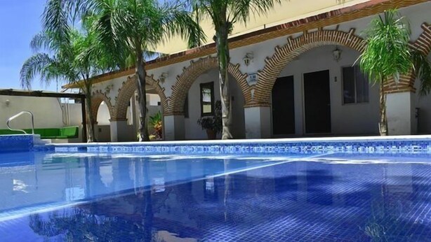Uma piscina com águas cristalinas azuis, rodeada por palmeiras e um edifício com portas em arco.