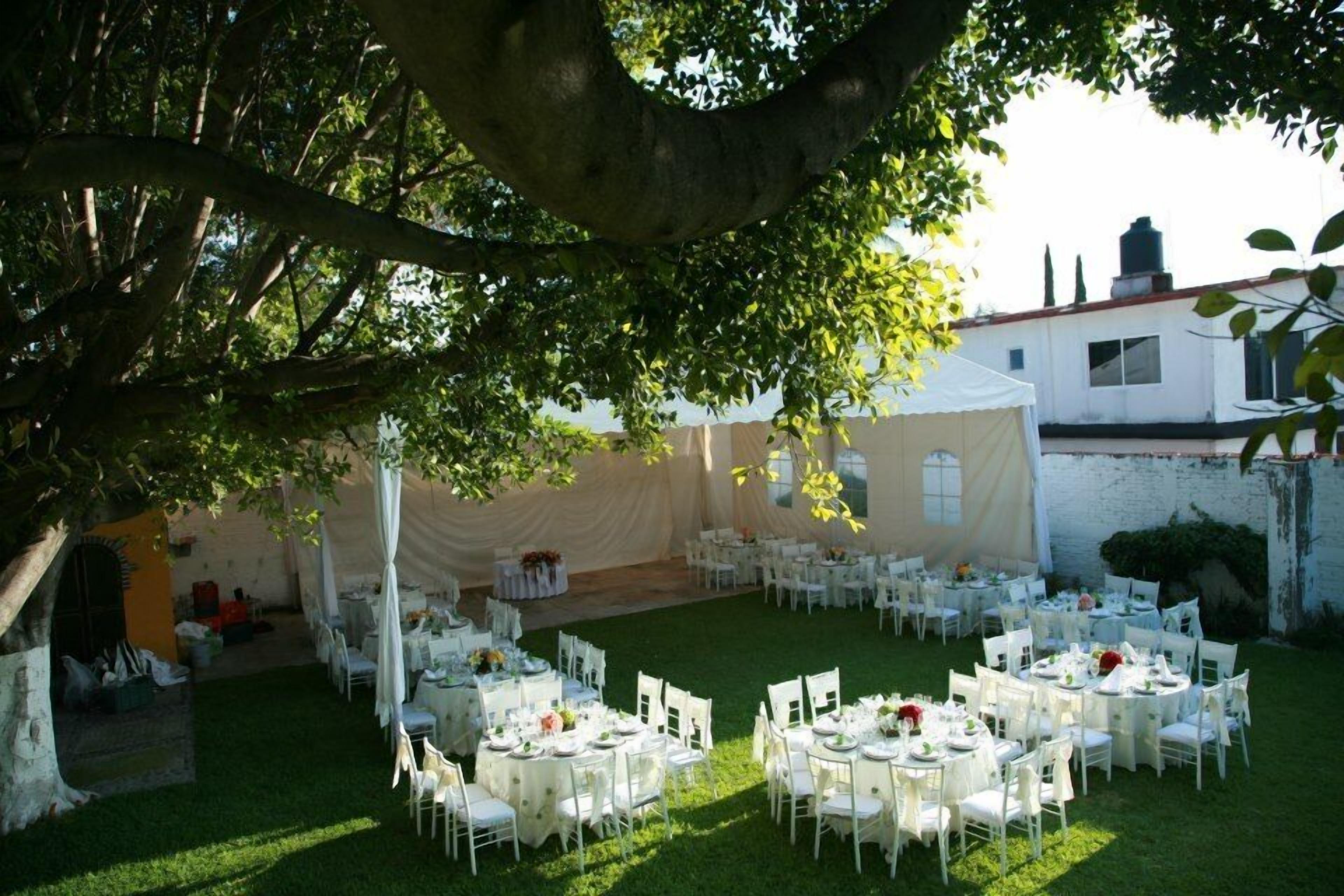 An outdoor wedding venue with round tables and white chairs, surrounded by a white tent and a tree.
