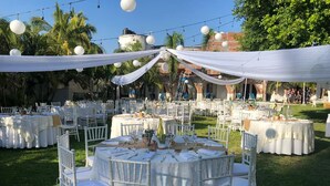 Uma recepção de casamento ao ar livre com mesas redondas, cadeiras brancas e luzes de fada.
