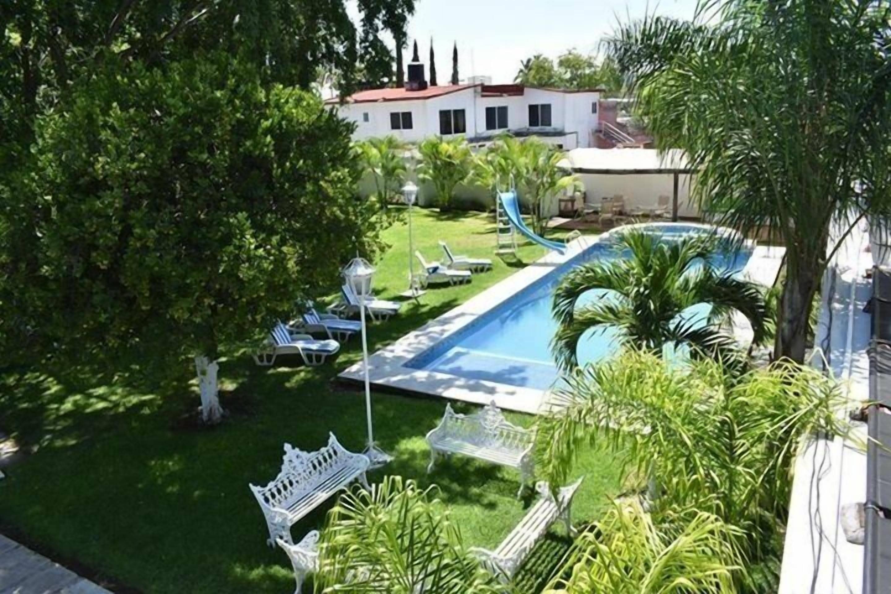 Ein Swimmingpool mit Rutsche, umgeben von einem gepflegten Rasen, weißen Bänken und Palmen.