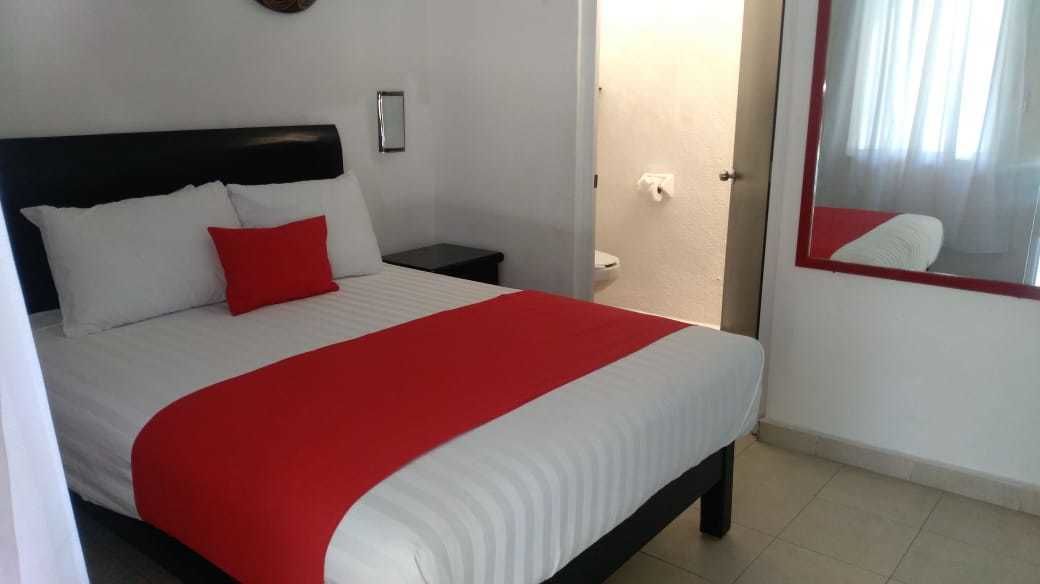 Una cama con ropa de cama blanca y roja, una mesita de noche, un espejo y una puerta que da a un baño.