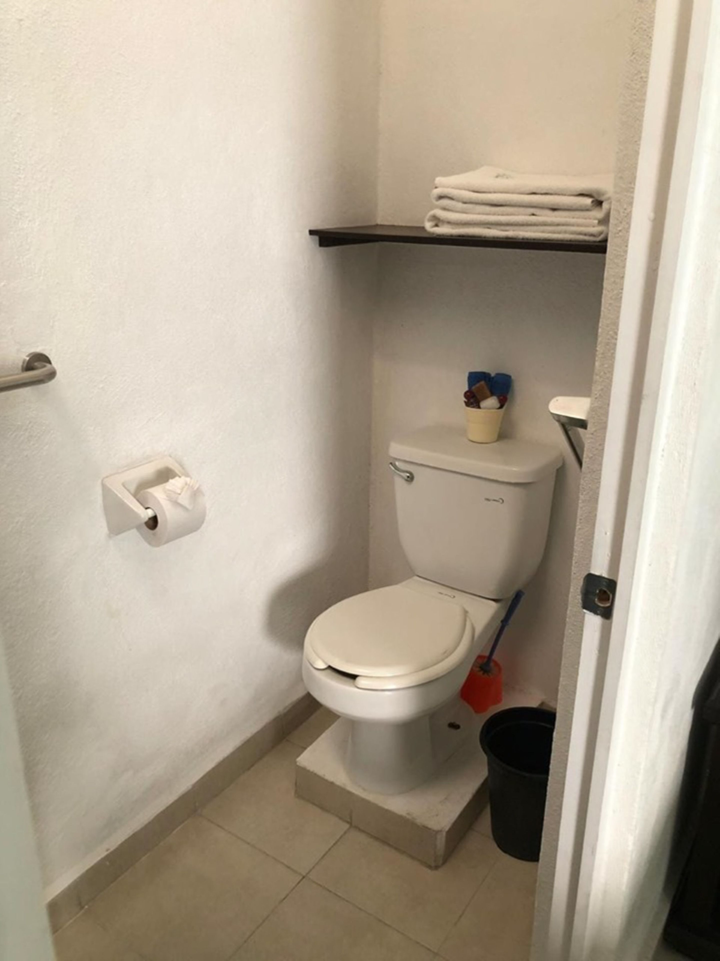 A small, clean bathroom with a toilet, a toilet paper holder, a trash bin, and a shelf with folded towels.