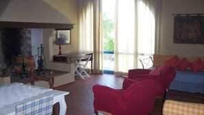 A living room with a fireplace, a dining table with a lace tablecloth, a red armchair, a sofa with checkered cushions, and a view of the outdoors through large windows. - Le Due Querce Agriturismo - Mimosa (Lazzeretto)