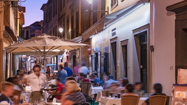 An outdoor dining area with tables and chairs, surrounded by buildings with shops and signs.