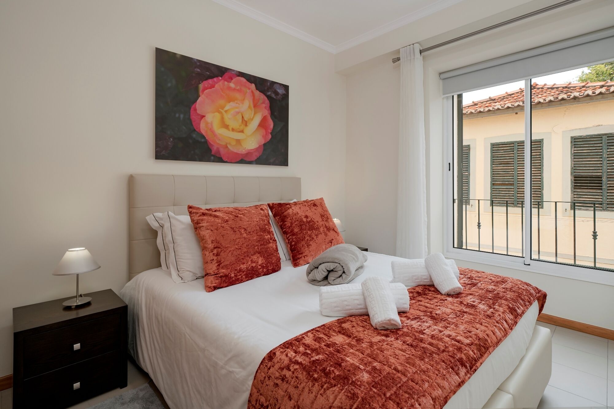 A neatly made bed with a white duvet and two red pillows, a bedside lamp, and a painting of a rose on the wall.