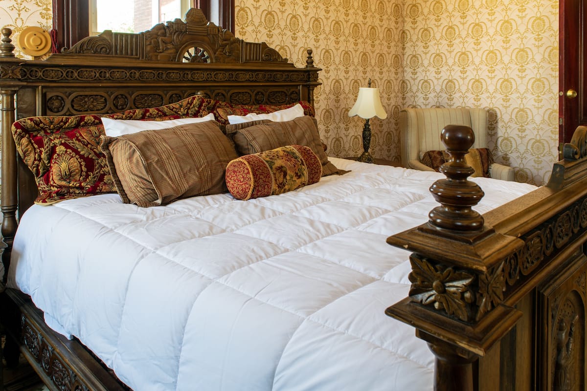 A neatly made bed with a white duvet and several decorative pillows, a wooden headboard, and a lamp on a bedside table.