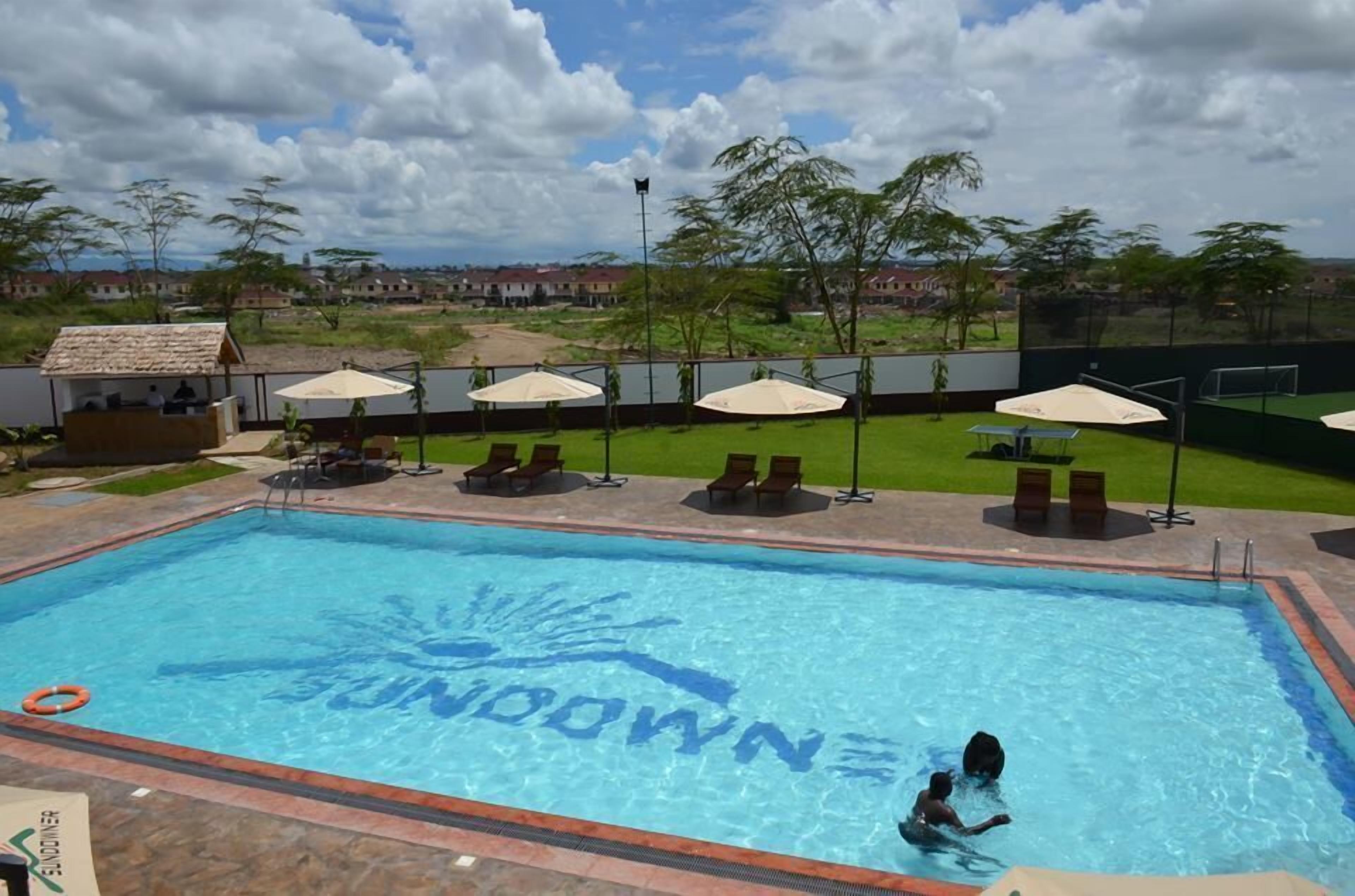 Una piscina con un diseño de sol, rodeada de tumbonas y sombrillas, con vista a casas al fondo.