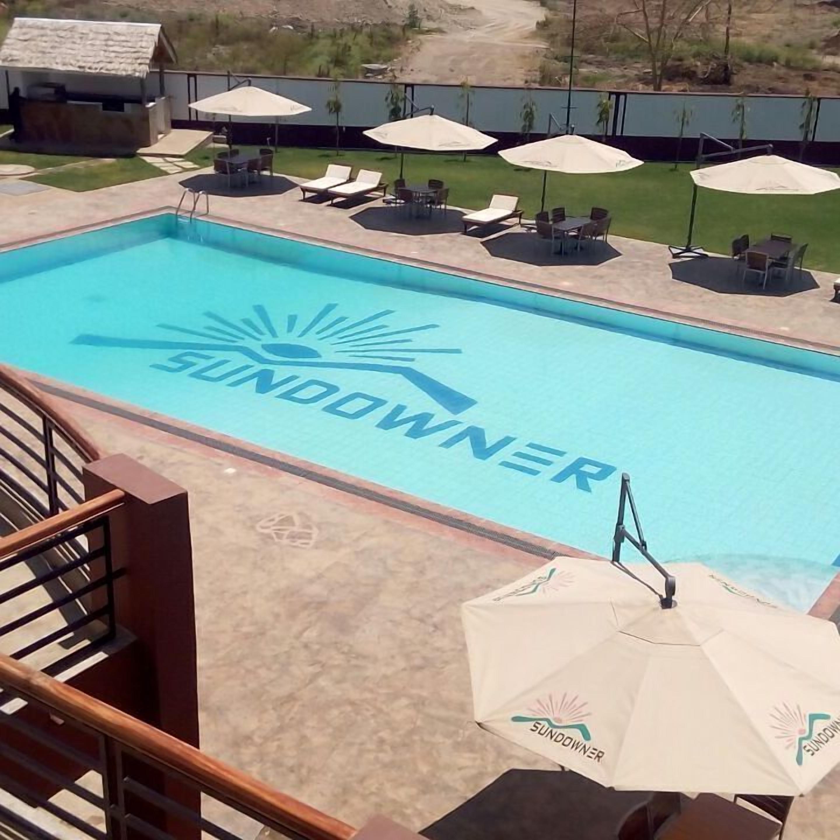 Una piscina con la palabra "SUNDOOWNER" escrita sobre el agua, rodeada de sillas de descanso y sombrillas.
