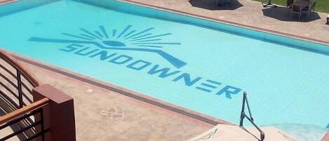A swimming pool with the word "SUNDOOWNER" on the water, surrounded by lounge chairs and umbrellas.