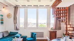 Ein gemütliches Zimmer mit einem Bett, einem blauen Sessel, einem Esstisch mit einem Metallkübel und Blick auf Gebäude durch große Fenster.