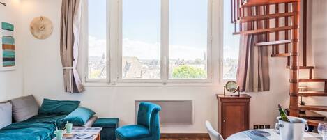 A cozy room with a bed, a blue chair, a dining table with a metal bucket, and a view of buildings through large windows.