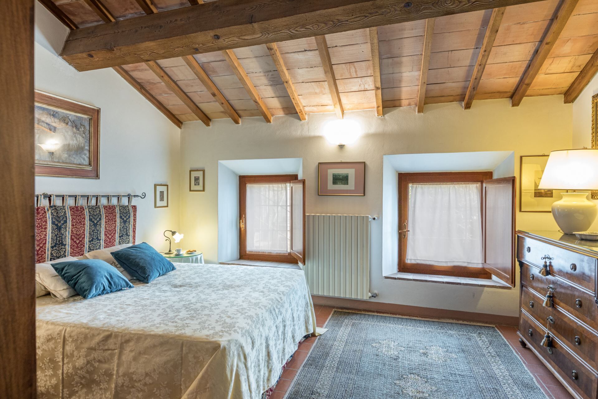 Une chambre à coucher comprenant un lit, une commode, deux fenêtres, un radiateur et un plafond avec des poutres apparentes.