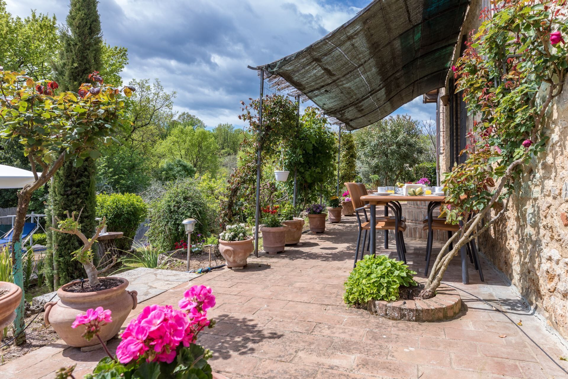 Un espace extérieur aménagé en terrasse, avec des jardinières en terre cuite, une table entourée de chaises et une pergola offrant de l’ombre.