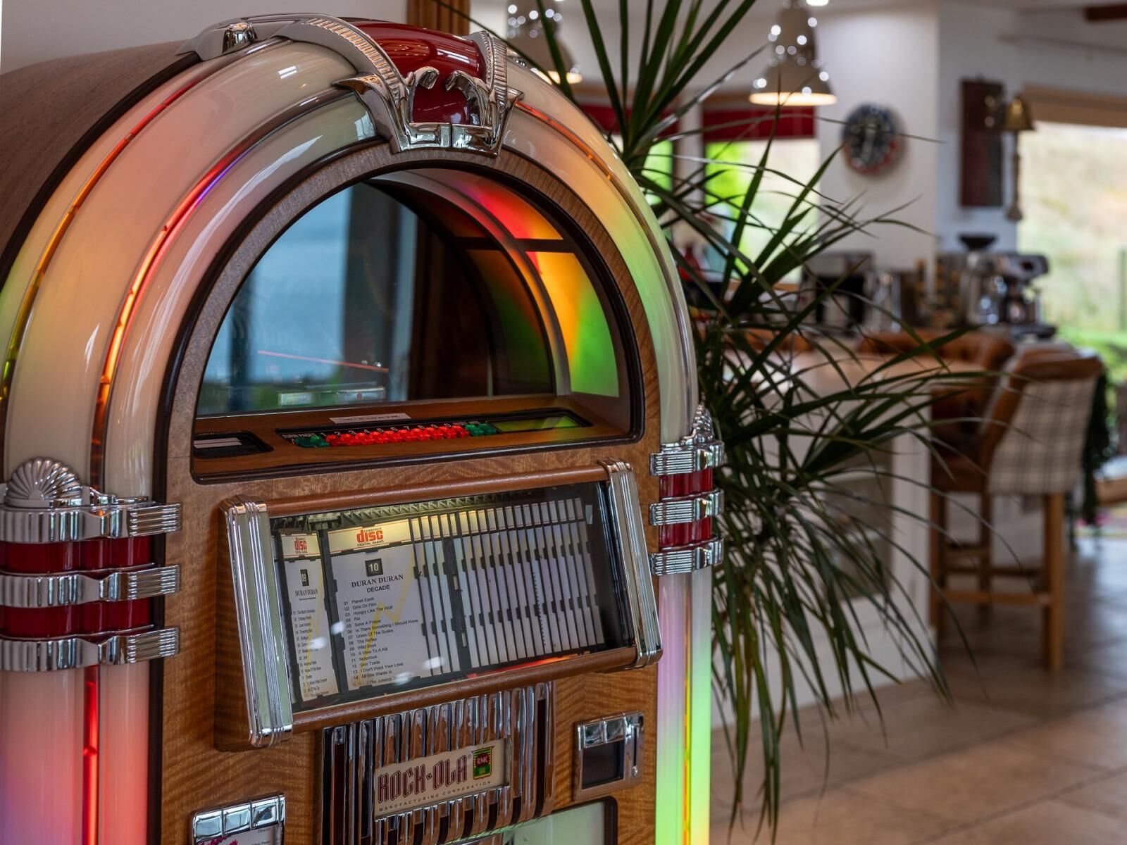Un juke-box au revêtement en bois et aux détails chromés, situé dans une pièce comprenant un coin repas et une plante.