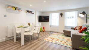 A modern living room with a dining area, a flat-screen TV, and a sofa with decorative pillows. - M02A - Modern 2Bed patio apartment in Hyde Park (London)