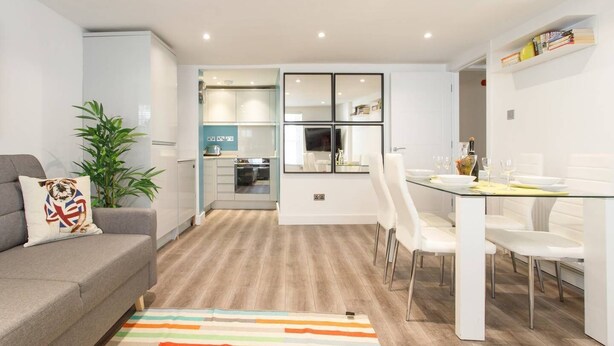 A modern living room with a grey sofa, a colorful striped rug, and a dining area with white chairs and a glass table. - M02A - Modern 2Bed patio apartment in Hyde Park (London)